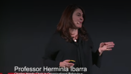 Female speaker on stage delivering a TEDx talk, with a headset mic and gesturing, while a lower-third bio identifies her as a professor of organizational behaviour.