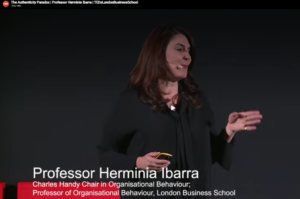 Female speaker on stage delivering a TEDx talk, with a headset mic and gesturing, while a lower-third bio identifies her as a professor of organizational behaviour.